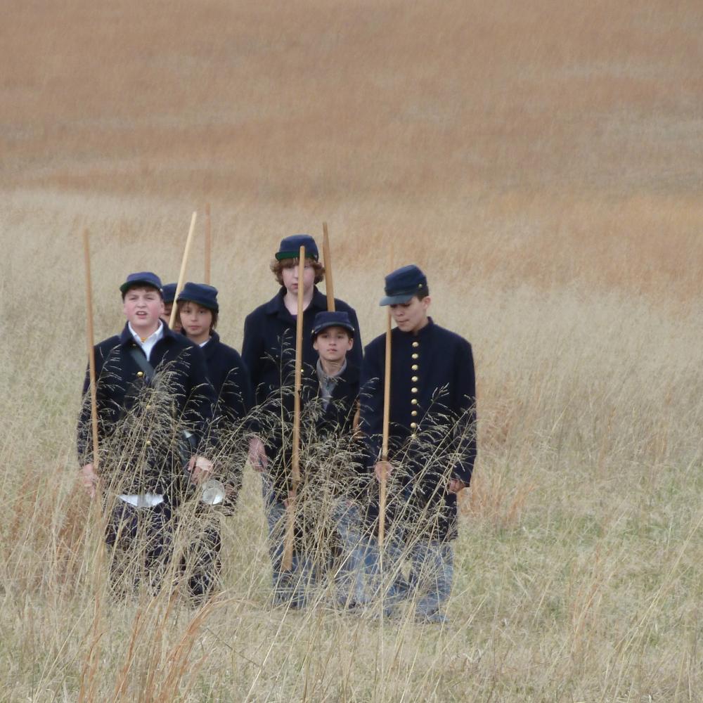 Color photo of a group of schoolboys re-enacting a Civil War march through a field. 