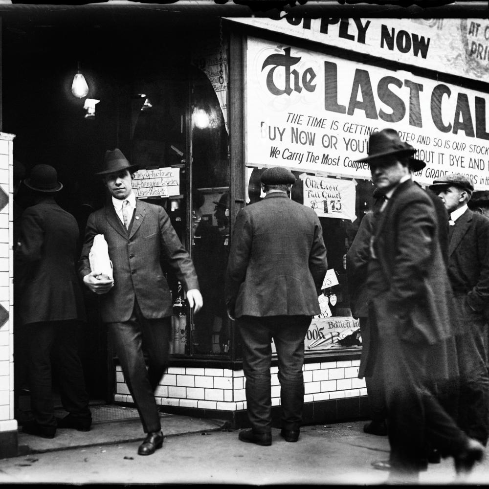 Black and white photo of men in formal attire heading into a liquor store. One man carries a packaged bottle in the foreground. 