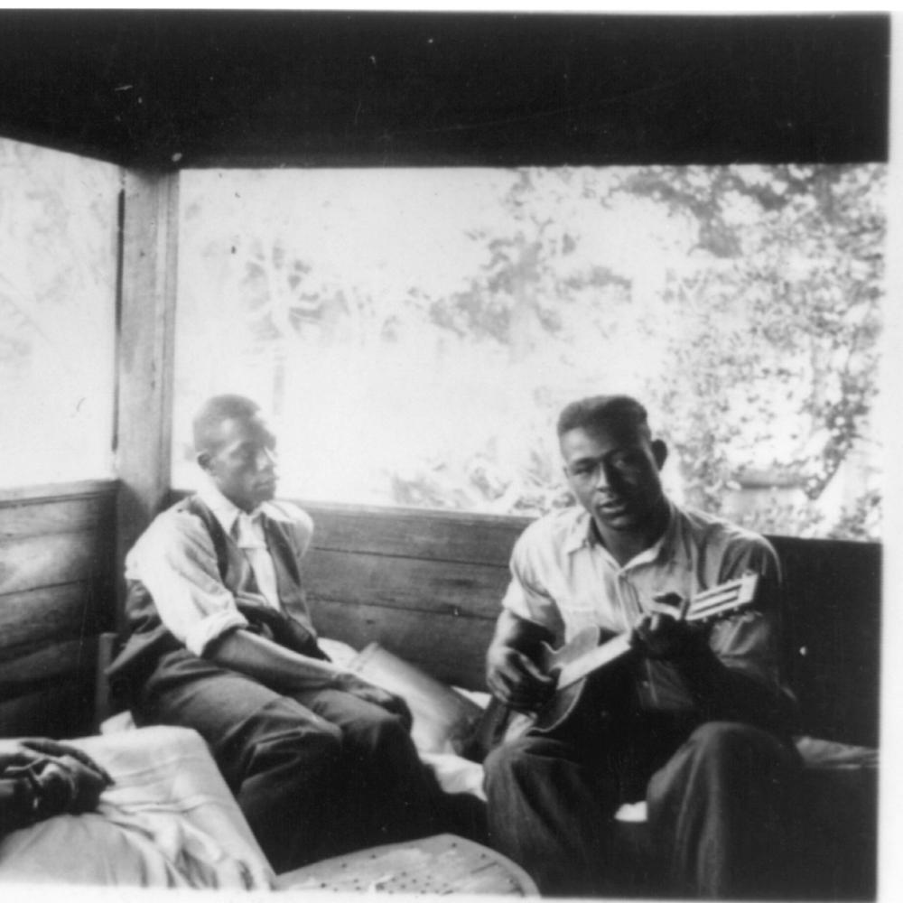 Black and white photo of three African Americans sitting on a porch, one of them playing the guitar.