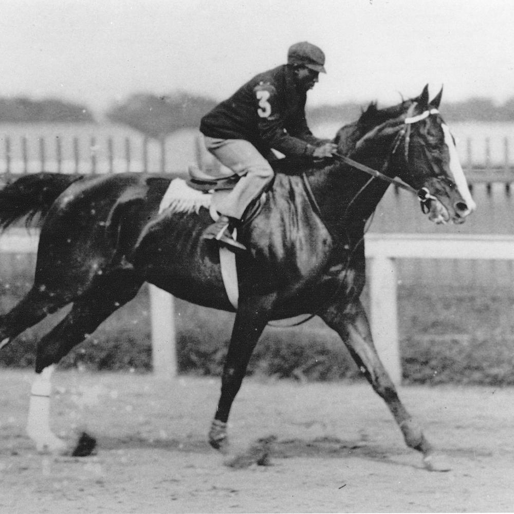 Black and white photo of an African-American jockey riding a horse at high speed. 