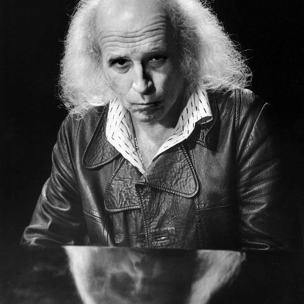 Black and white photo of an aged musician, slightly balded, with long, white, frizzy hair. He wears a leather jacket and collared shirt underneath.