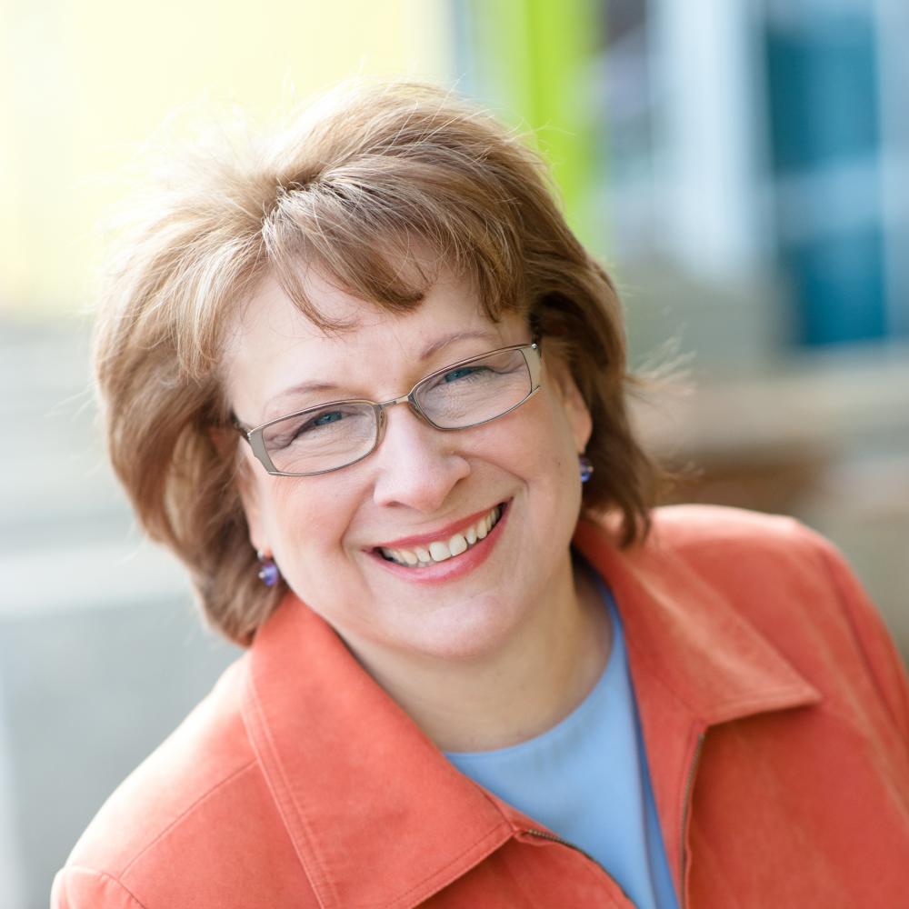 Color photo portrait of Maggie Coval in a long-sleeved orange blazer.