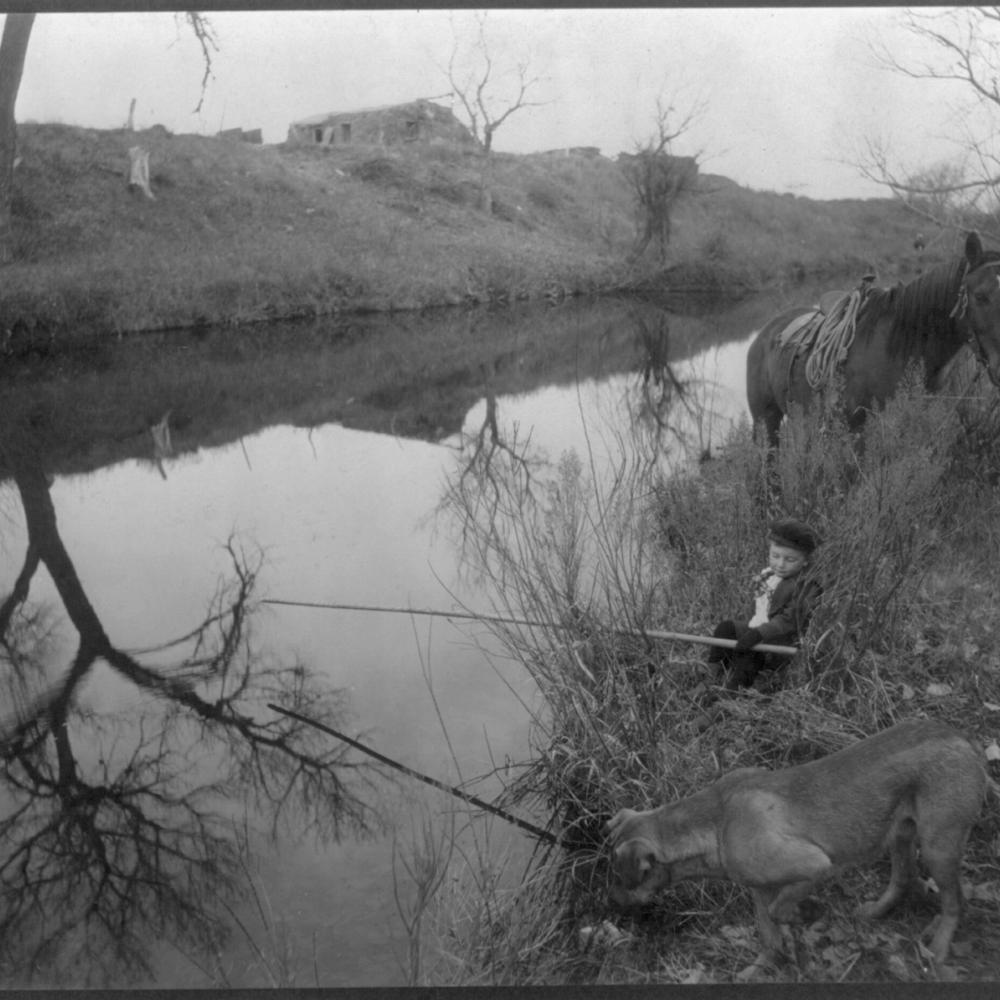 Black and white photo of a little boy fishing in a stream while his dog attempts the same maneuver in the foreground.