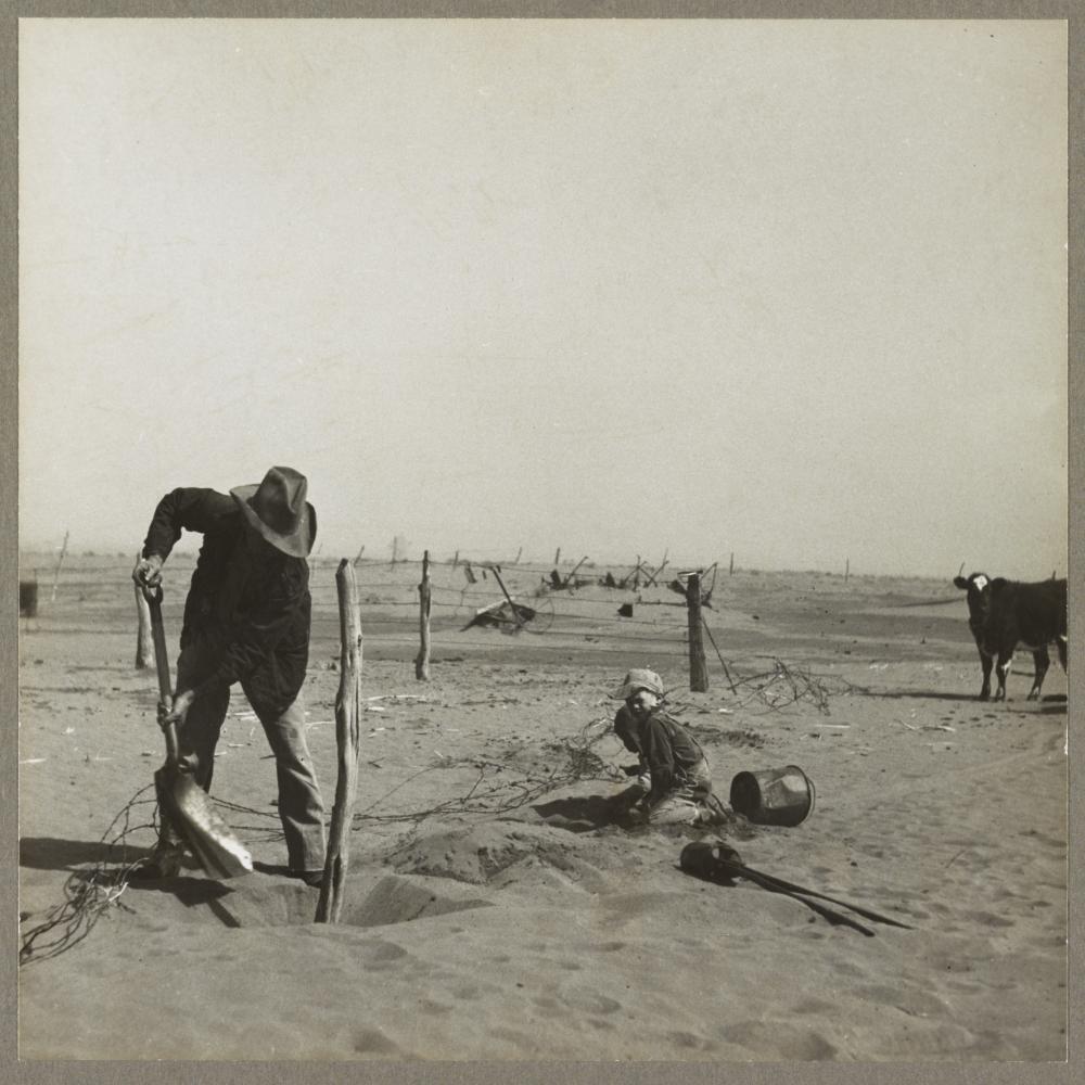 Black and white photo of a man shoveling sand in the foreground, with a child to his right, sitting on the plain, and a cow further in the distance.