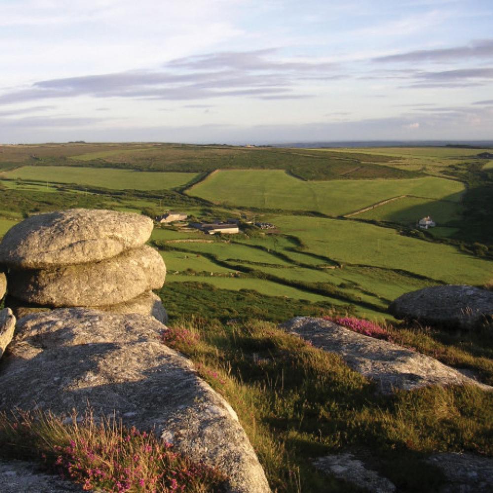 Lush green farmland extends into the distance below a hill covered in stones and grass, with late afternoon light