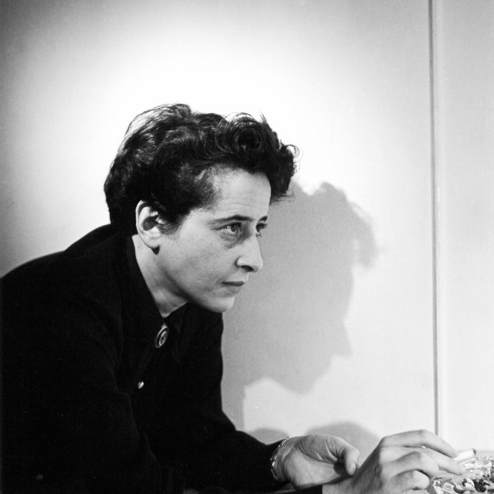 Arendt, with short dark hair, holding a cigarette, leaning on a countertop and facing to the right