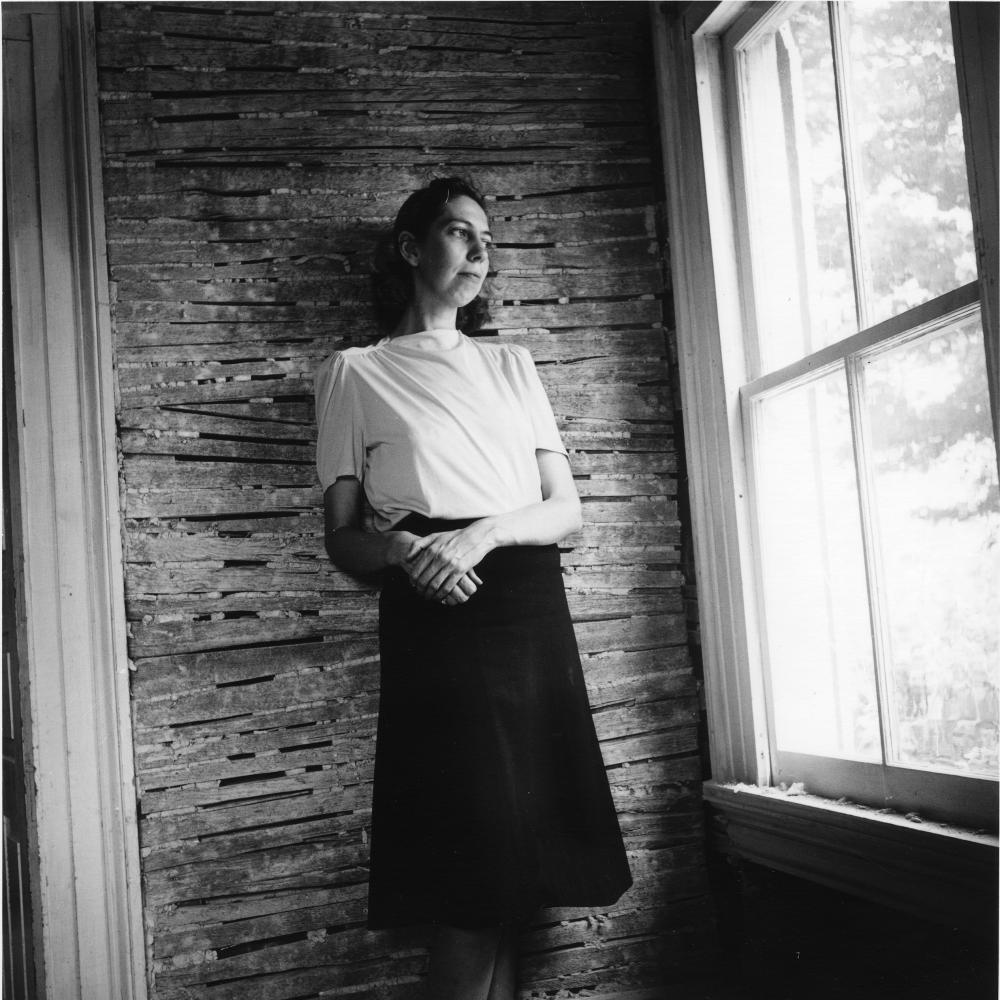 Welty in a white blouse and black skirt, leaning against a brick wall and looking out of a large window to her left