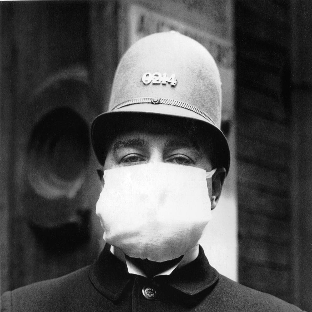 A police officer, in uniform and rounded helmet, has his entire face, other than the eyes, covered by a white cotton medical mask