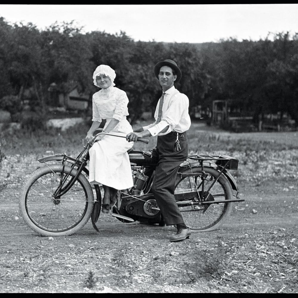 Kratzer, in a white shirt and black hat, sits on a motorcycle behind a woman in a white dress and cap, on a dirt road