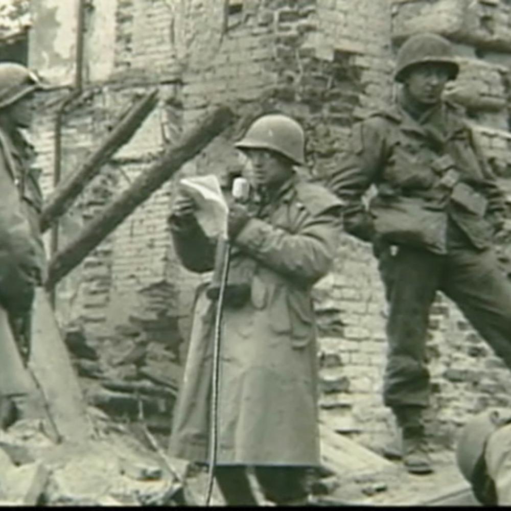 Three armed and uniformed soldiers watch Plambeck, also uniformed, speak into a microphone while reading off of a paper, in the ruins of a stone building