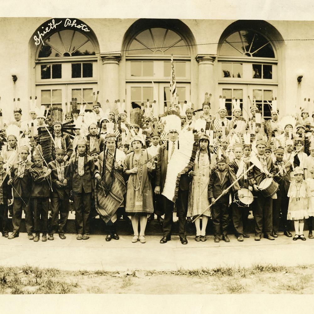 Sepia-colored photograph of a tribe of Native Americans standing in front of a large building.