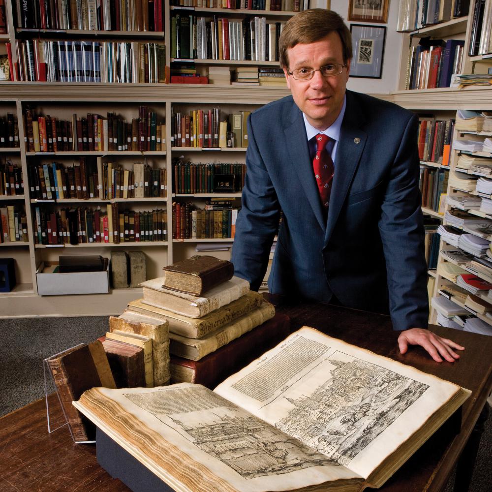 Photo of a man in a suit leaning over a rare book in a library, while looking at the camera.