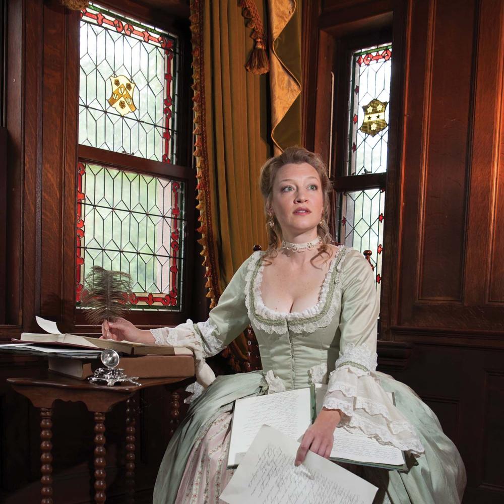 Stauffer in a green, 1800's era dress, sitting at a desk