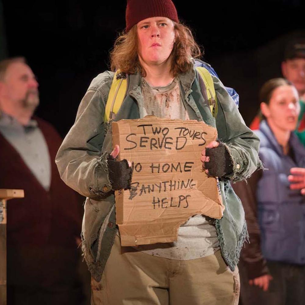 An actor holds up a sign reading "served two tours, no home, anything helps" during a scene