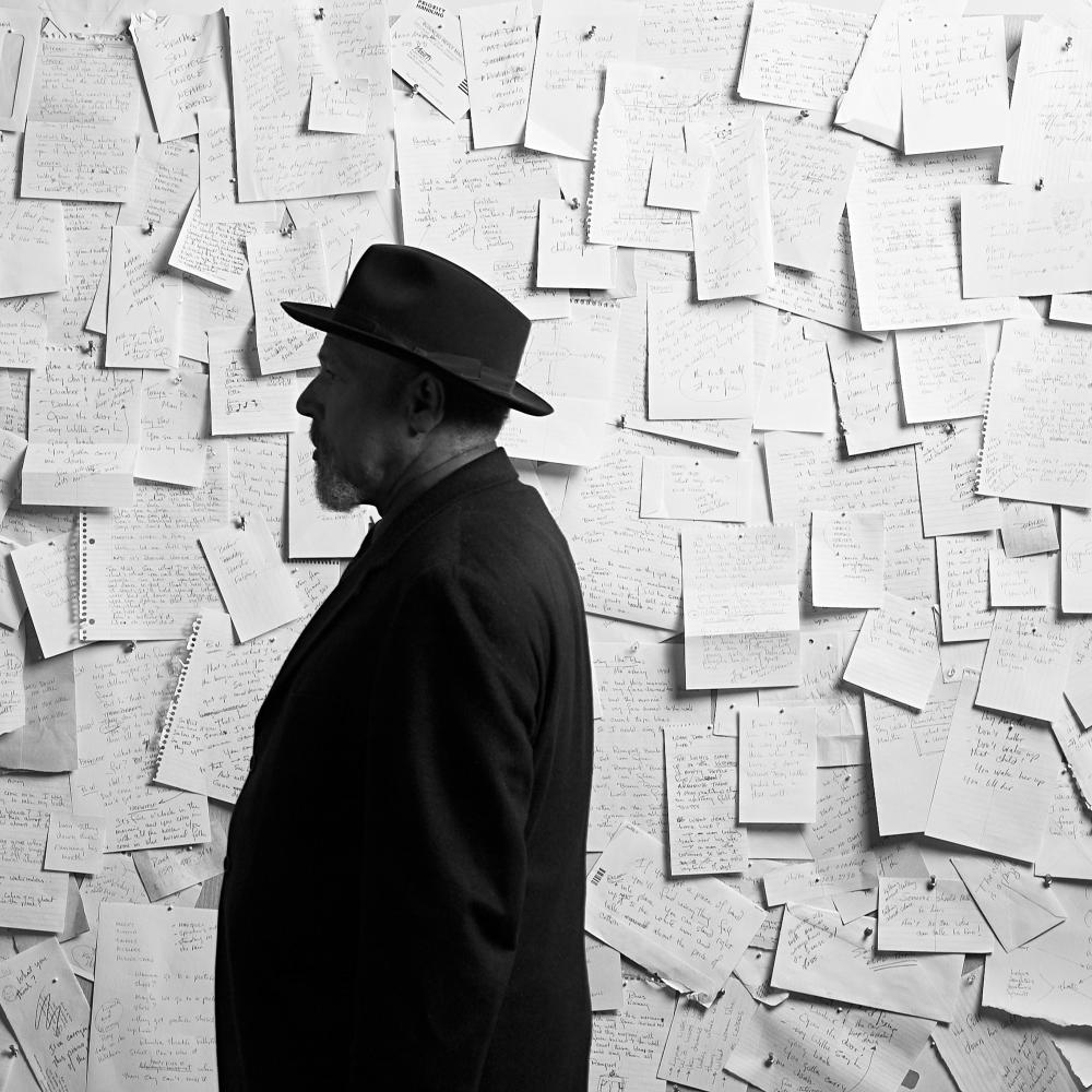 Black and white side portrait of August Wilson standing before a wall covered by letters that have been pinned onto it.