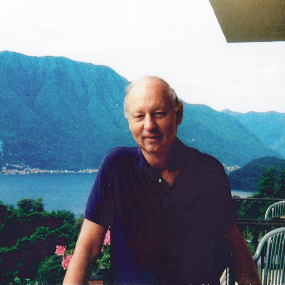 Color photo of Jules Witcover standing on the balcony of a house which overlooks a large bay and mountains in the background.