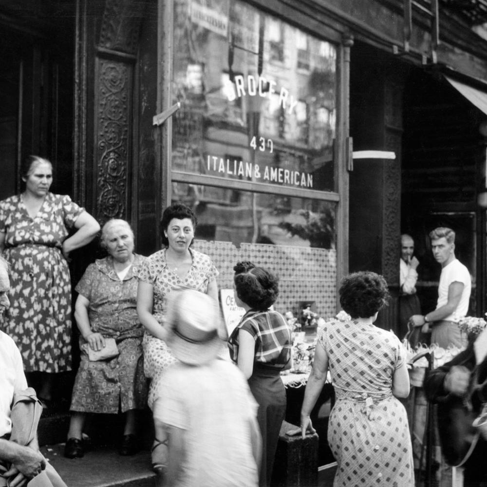 Black and white photo of Italian immigrants in Little Italy congregating outside a storefront. 