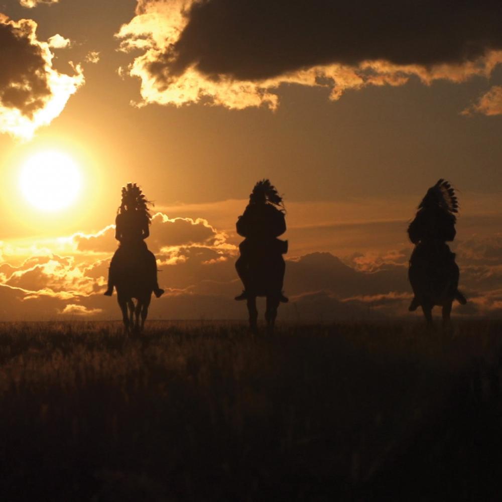 Color photo of three men on horseback riding into the glimmering sunset.