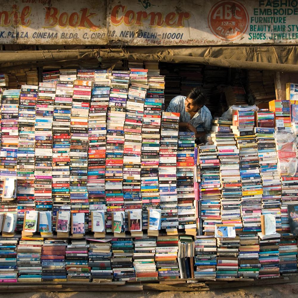 Color photo of a man in Delhi, India, peering out from behind a wall of books which comprises his storefront.