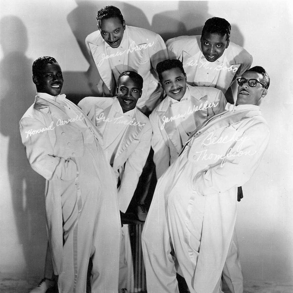 Black and white photo of a musical group wearing white tuxedos and posing for a photograph at irregular angles.