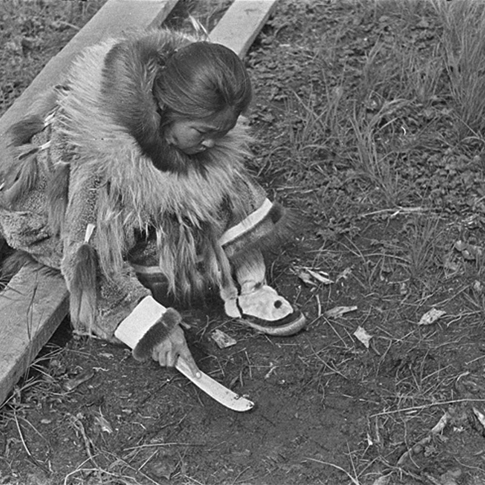 Black and white photo of an Indian woman sitting on the ground with a knife-shaped tool in her hand.