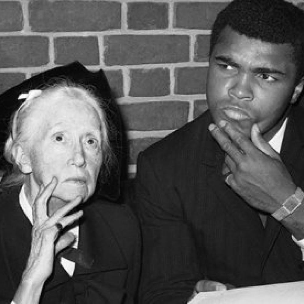 Black and white photo of poet Marianne Moore seated next to Mohammed Ali, who stares, intrigued, fingers on his chin, toward something else in the room.
