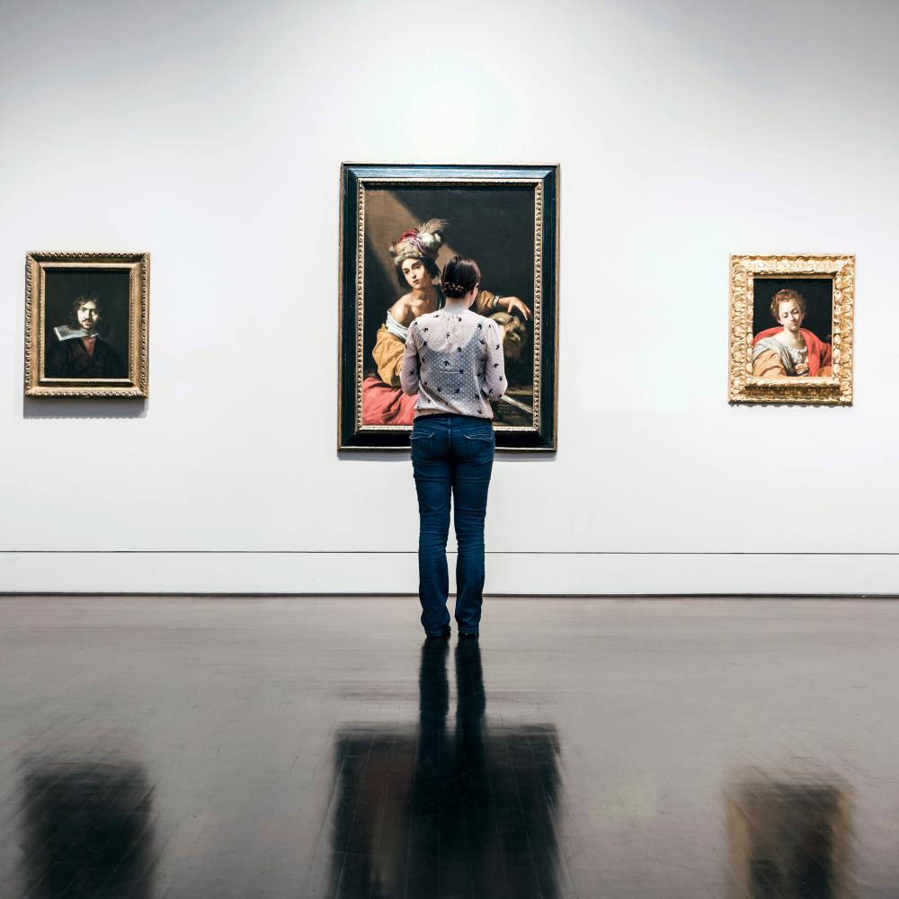 Photograph of woman standing in museum, three paintings on the wall