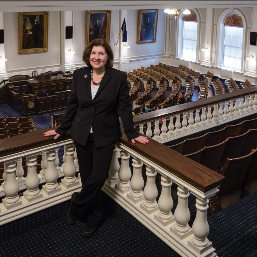 Color photo of Deborah Watrous standing in the gallery of the New Hampshire State House. The seats of representatives can be seen below her. 