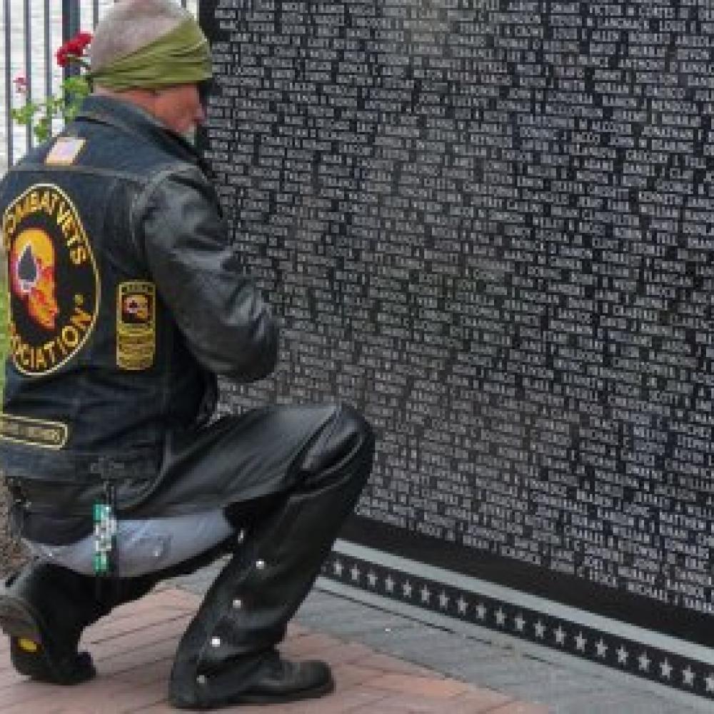 A man in jeans and a motorcycle jacket kneels before the Vietnam War Memorial.