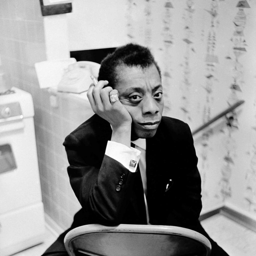 Black and white photo of James Baldwin, sitting backwards on a foldable chair, in a kitchen. He's is wearing a black suit with a white shirt and cufflinks. He is holding a cigarette in his right hand with a bold pink ring.