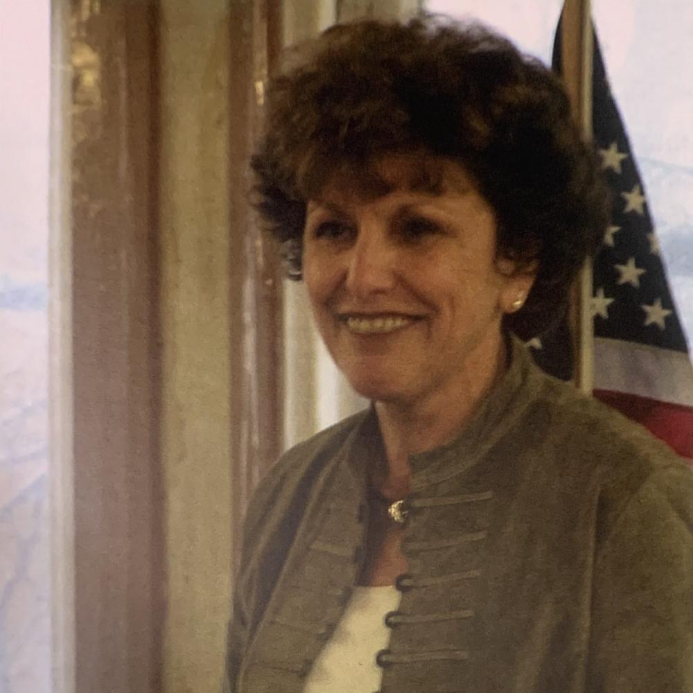 Portrait of Jan Fedewa standing at a podium with a US flag in the background. She is in a light brown, unbutton shirt jacket with a white shirt. 