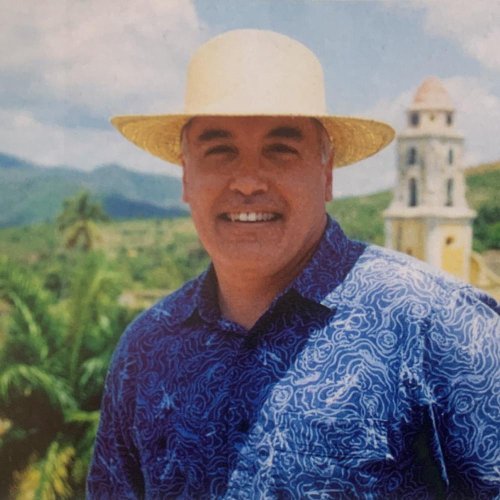 Portrait of David Tebaldi in a blue, decorative shirt and straw hat., with a countryside background.