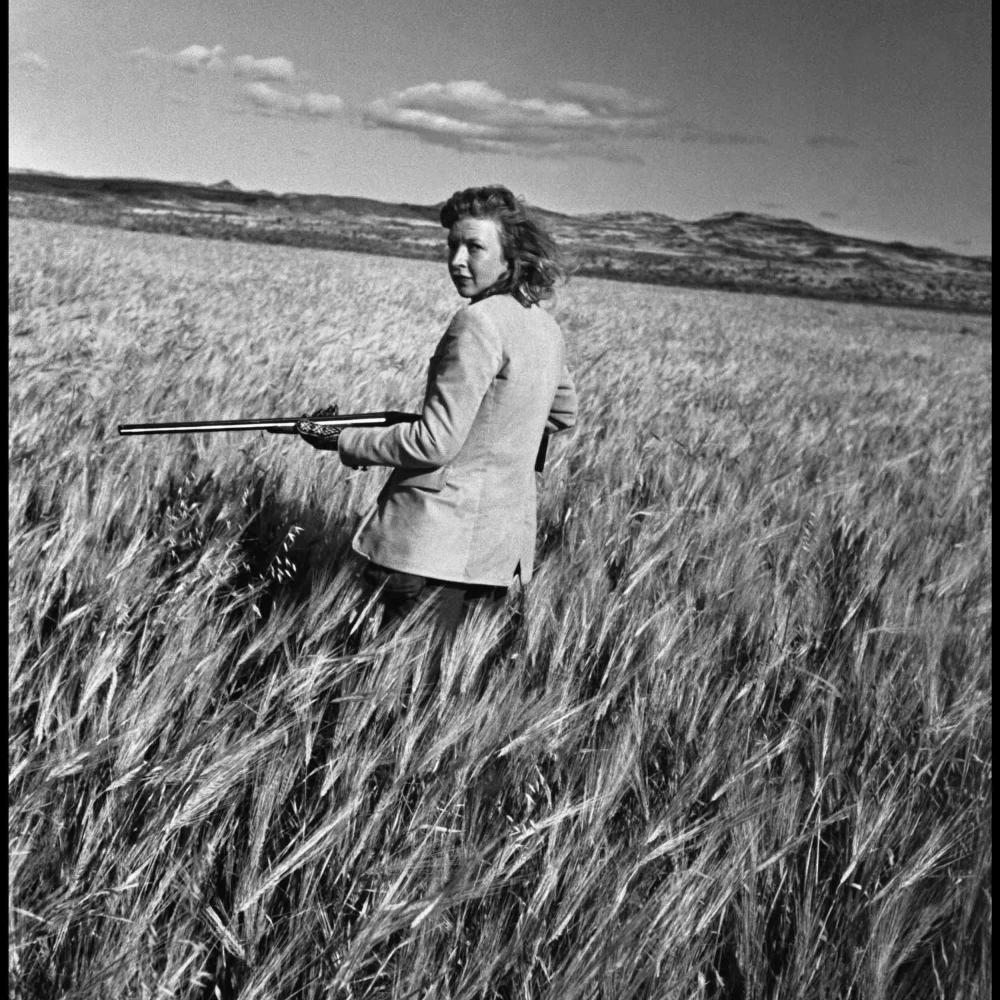 Martha Gellhorn toting rifle or shotgun in field.