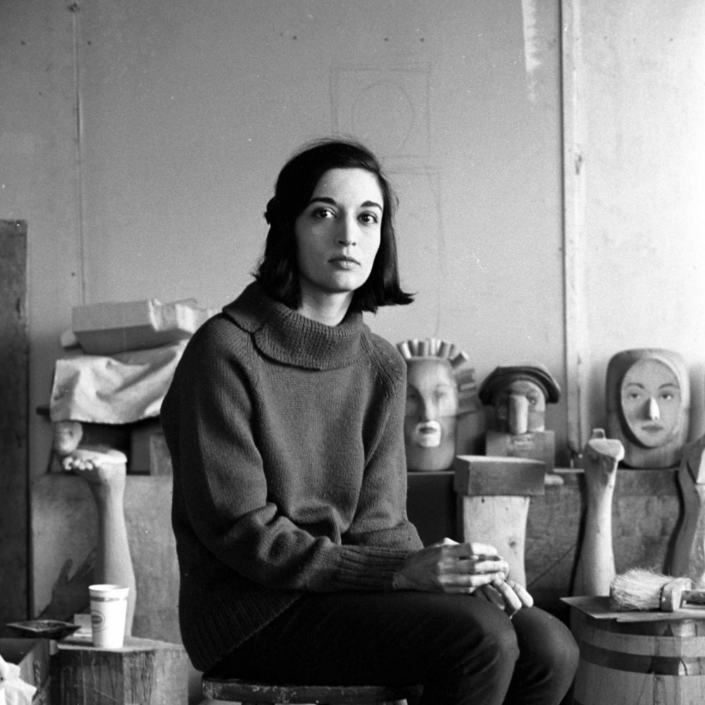 Marisol Escobar seated on stool with pieces of artwork behind her. 