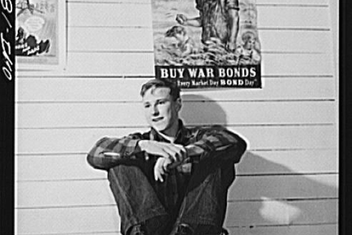 Black and white photo of young man sitting on table, war poster behind him