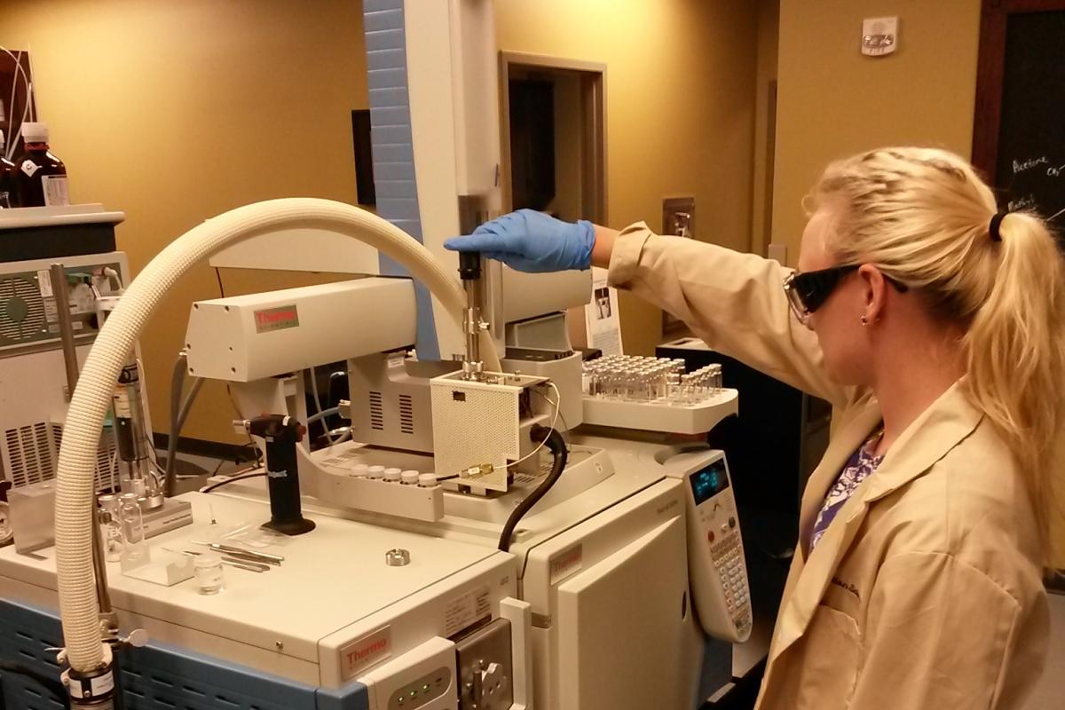 Woman stands at machine in lab coat and goggles