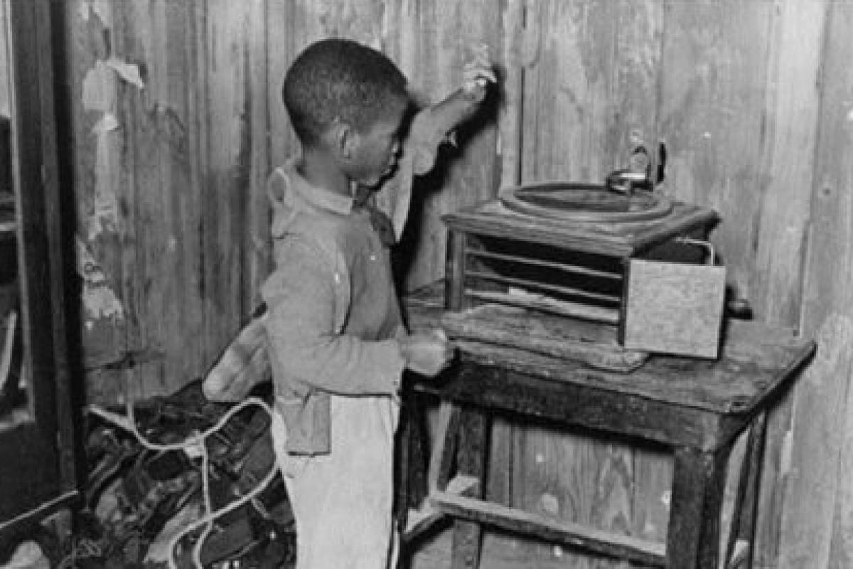 Black and white photo of a little black child playing with a phonograph in a cabin.