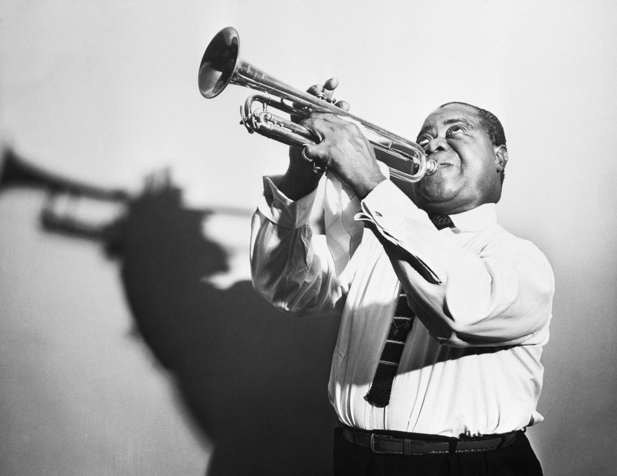 Armstrong in a white collared shirt and tie, holds his cornet in the air as he plays