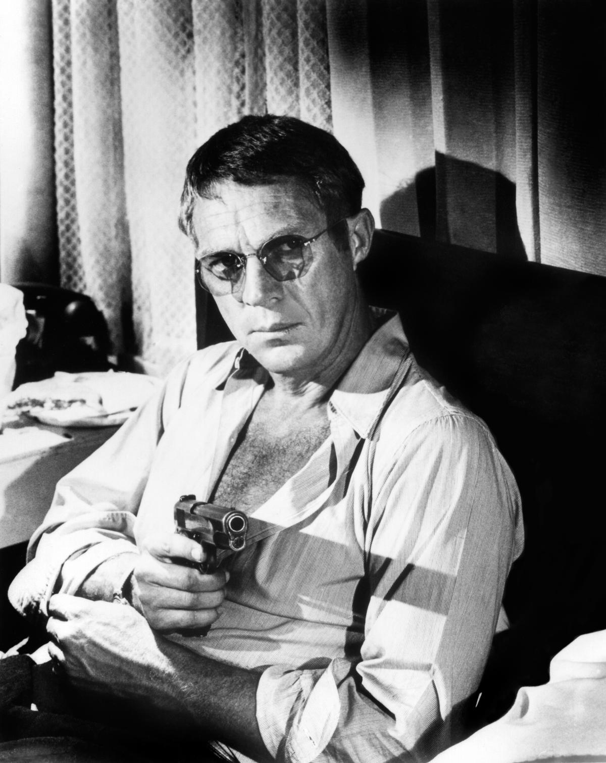 McQueen, wearing tinted sunglasses and a shirt half-unbuttoned, casually points a gun
