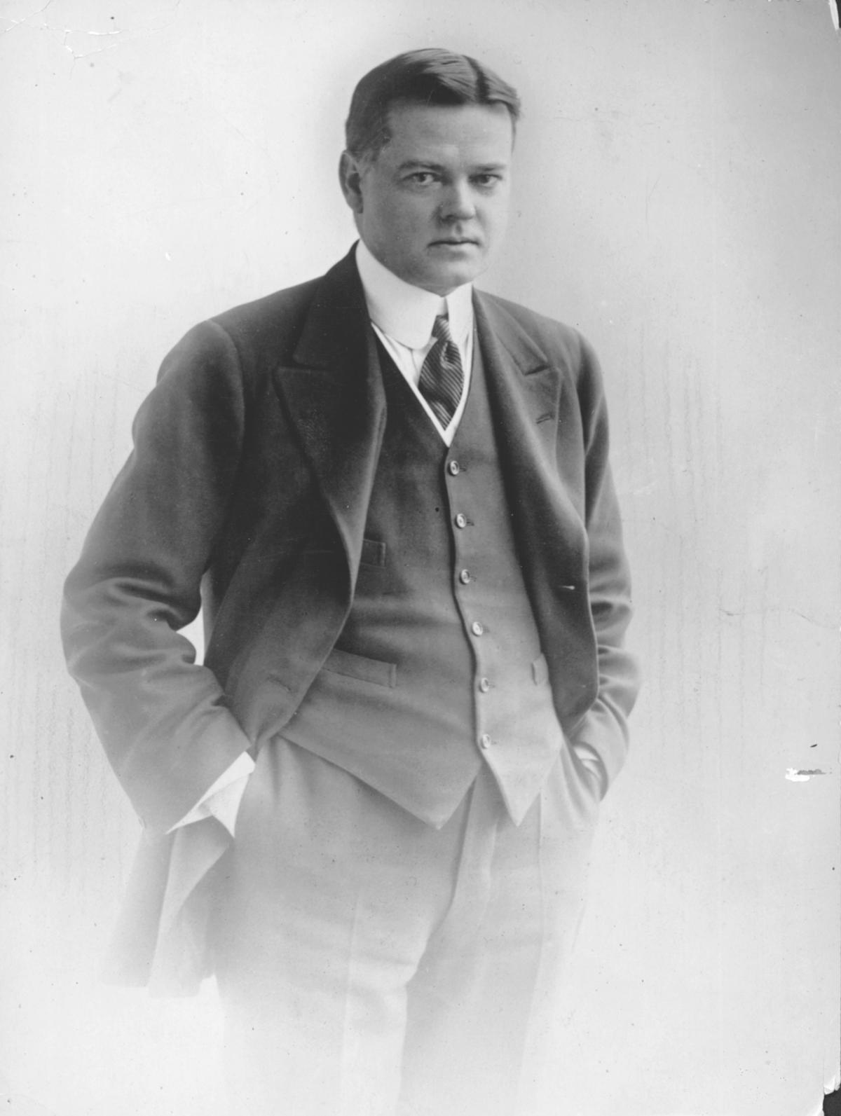 Hoover standing with hands in pockets, wearing a dark suit, hair slicked back