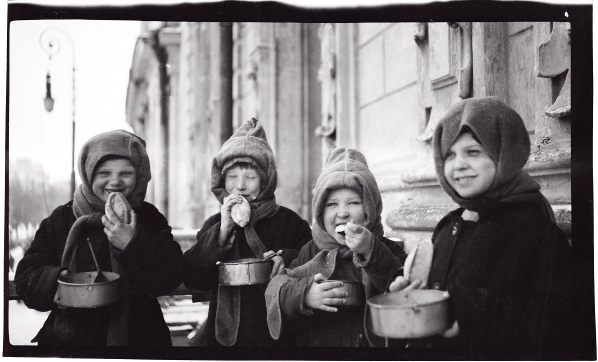 Four girls in winter coats and hoods eat bread and hold tin bowls of food
