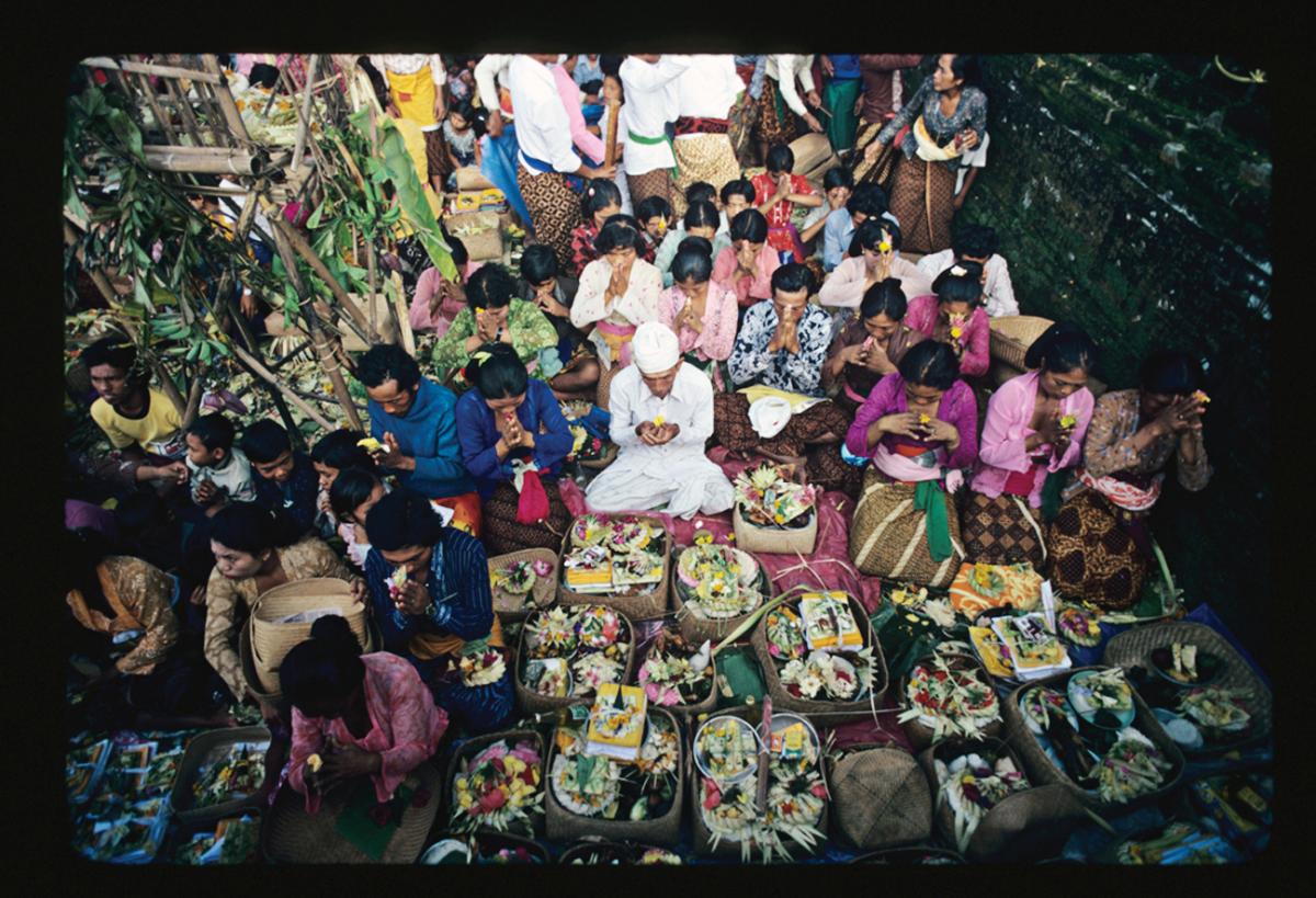 A crowd of worshippers sit with palms clasped, while a priest in white leads prayer and presents their floral offerings to the gods