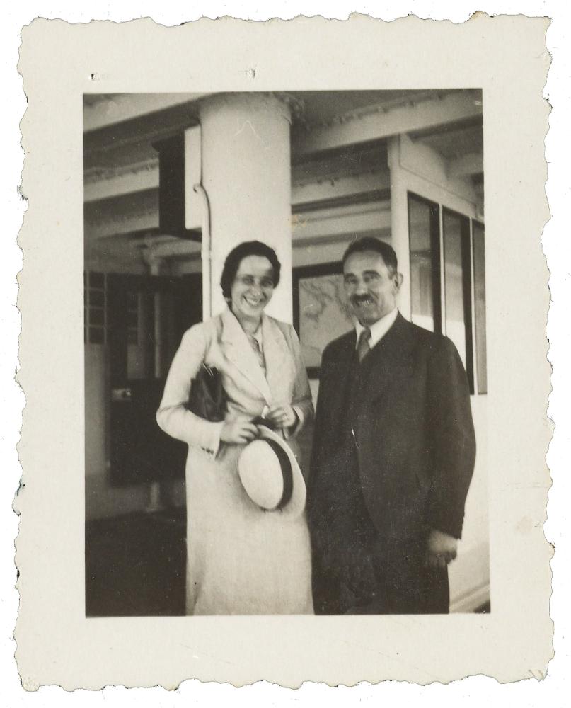 Arendt, smiling and wearing a long white coat, holding a hat, stands next to a moustached man in a dark suit