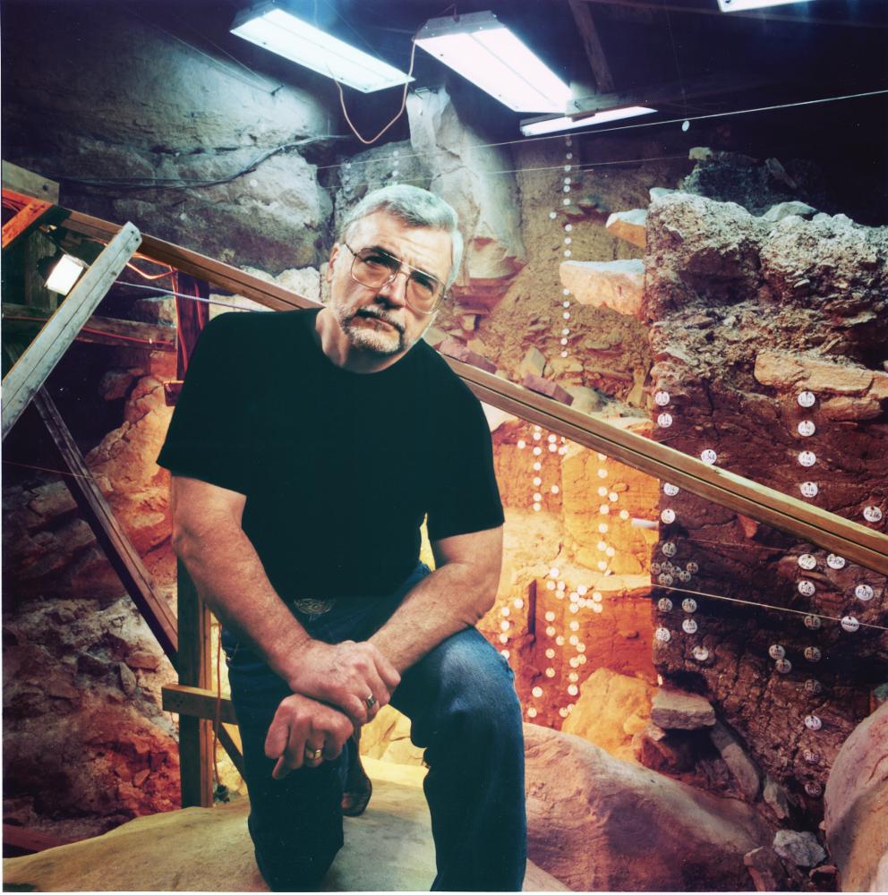 Adovasio kneels inside of an excavation site, filled with rock formations and wooden scaffolding