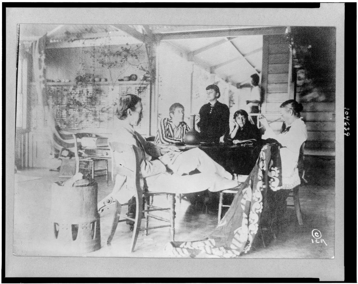 stevenson in the foreground, sitting and resting his legs on a chair, while his children sit at the table with him