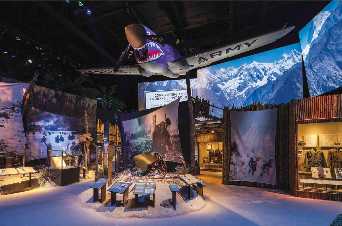 A shark-faced P-40 Warhawk is hanging on the ceiling of "The Road to Tokyo" exhibit at the National World War II Museum. There are other imagery of soliders, palm trees, and wooden street signs. 