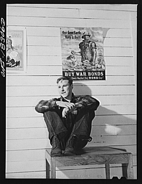 Black and white photo of young man sitting on table, war poster behind him
