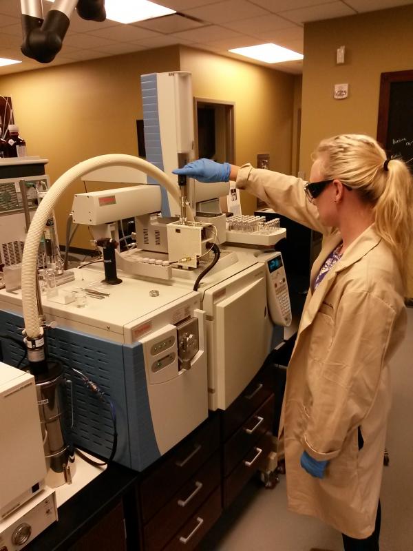 Woman stands at machine in lab coat and goggles