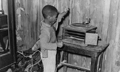 Black and white photo of a little black child playing with a phonograph in a cabin.