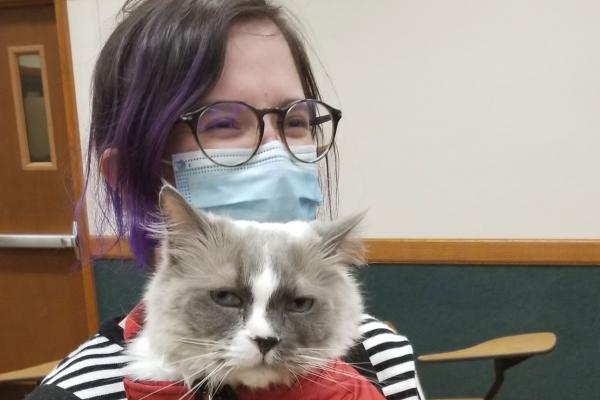 A laughing woman with purple hair carrying a glaring cat in a puffy vest.